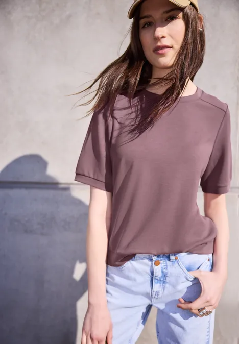 Person vor grauer Wand in mauvefarbenem T-Shirt und hellen Jeans, Hand in Tasche.